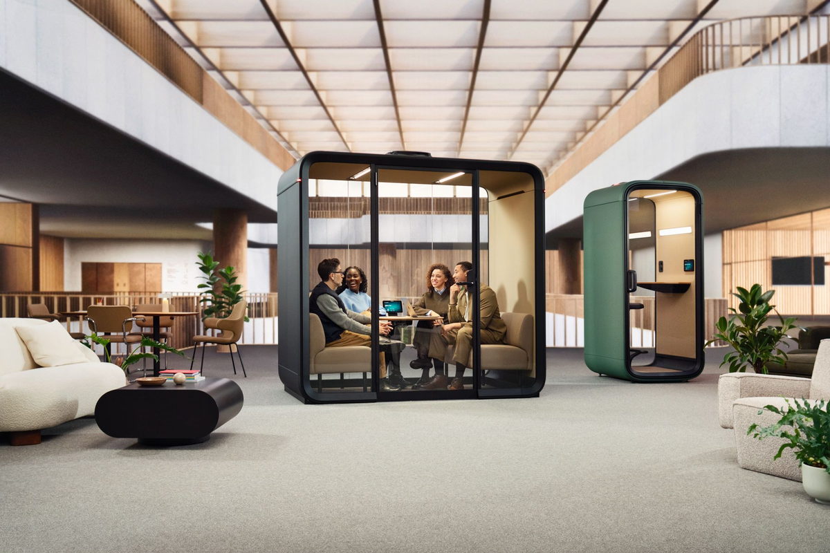 Cuatro personas reunidas en una cabina acústica de cristal y una cabina individual verde al fondo, en el atrio de un edificio de oficinas